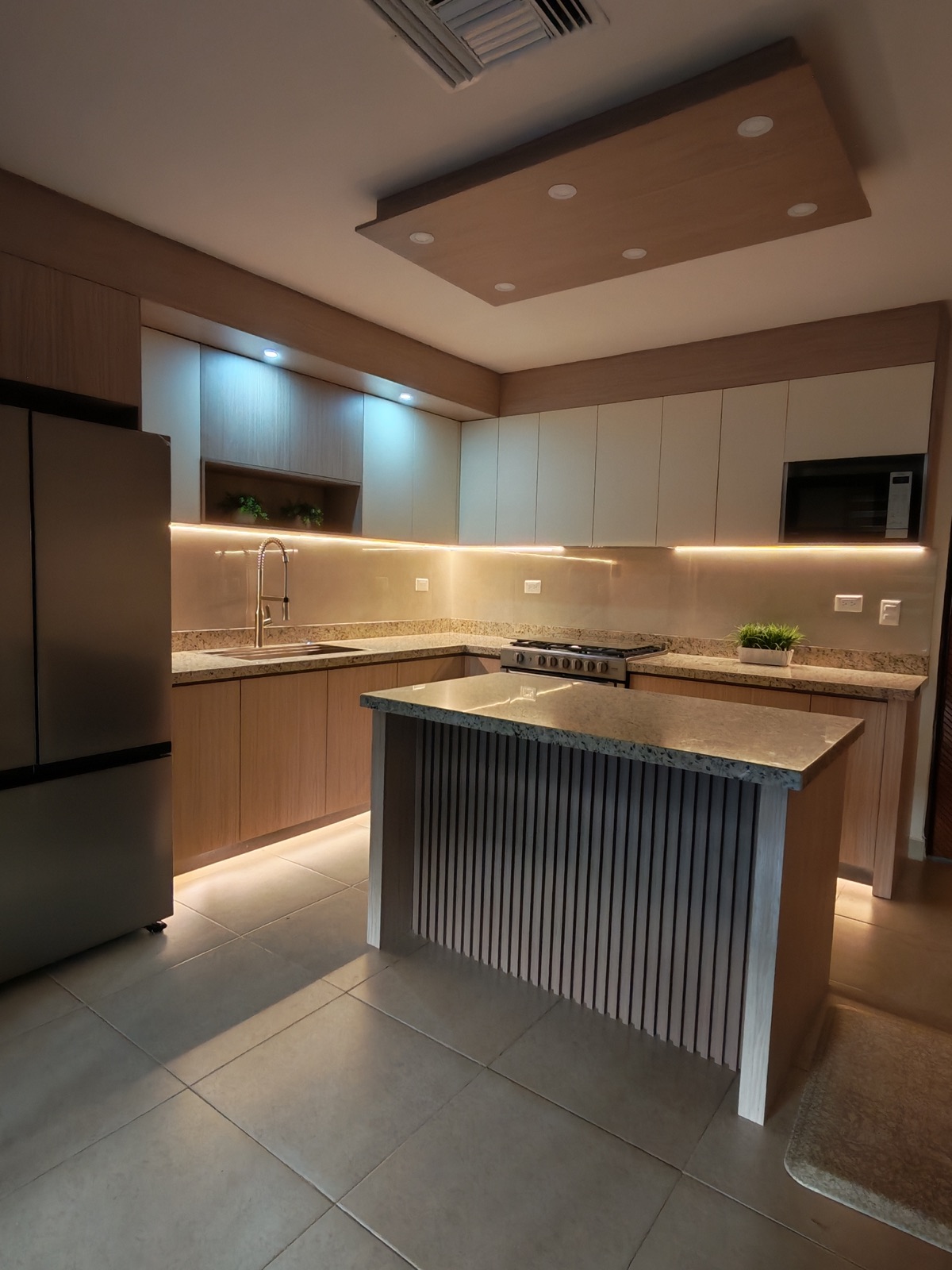 Cocina de noche con iluminación LED bajo gabinetes, plafón de madera e isla de granito